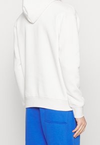 Felpa bianca con cappuccio e tessuto morbido, presenta una tasca a marsupio e polsini a costine, abbinata a pantaloni della tuta blu.