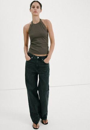 Femeie purtând un top fără mâneci, de culoare verde măsliniu închis, cu dantelă la margini și blugi negri largi, stând cu mâinile în buzunare în fața unui fundal alb simplu.