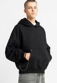 Czarna bluza z kapturem na sznurki, o luźnym kroju, z prążkowanym dołem oraz boczną kieszenią ozdobioną srebrnym łańcuszkiem. Wykonana z miękkiej, teksturowanej tkaniny.