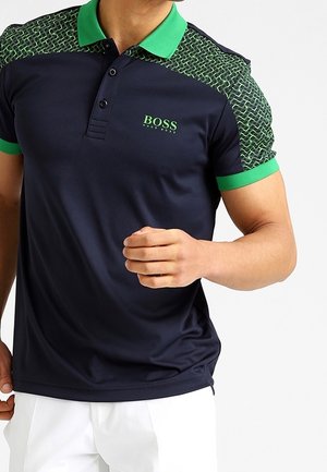 Homme portant un polo bleu marine avec col vert, épaules à motifs verts et noirs, poignets de manches verts, et logo "BOSS Hugo Boss" sur la poitrine.