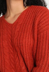 Pull en maille rouge avec un col en V, présentant un motif torsadé texturé et un tissu à tissage lâche. Accentué par une couture visible le long de l'encolure.