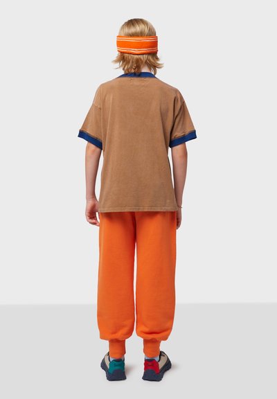 Braunes T-Shirt mit marineblauen Ärmeln, orangene Sweatpants und schwarze Sneakers mit türkisfarbenen und roten Akzenten; Stirnband mit orangefarbenen Streifen.