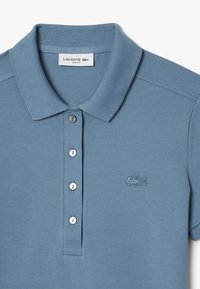 Vaaleansininen polo-paita, joka on valmistettu teksturoidusta kankaasta. Siinä on kaksinappinen suljenta, normaali kaulus ja brodeerattu logo vasemmassa rinnassa.