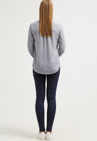 Camicia a maniche lunghe blu con motivo, con orlo arrotondato, abbinata a jeans skinny scuri e sneakers bianche, vista da dietro.