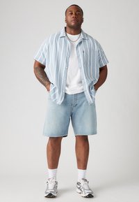 Chemise à manches courtes, à rayures bleu clair, sur un T-shirt blanc, associée à un short en denim léger et des chaussures de sport grises.