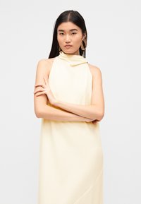 Κίτρινο φόρεμα με halter λαιμόκοψη, με ντραπέ ντεκολτέ, λεία υφή και με διαγώνια ραφή ως διακοσμητικό στοιχείο. Το μοντέλο φοράει μεγάλα χρυσά σκουλαρίκια κρίκους.