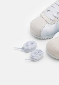 Sneaker grigio chiaro con punta in scamosciato e corpo in tessuto liscio, accompagnata da due lacci azzurri arrotolati.