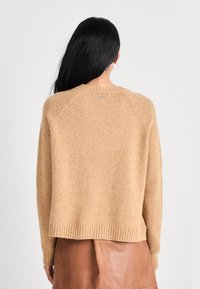 Pull en tricot beige texturé avec un col rond, un ourlet côtelé et des manches raglan. Présente un petit logo au col arrière.