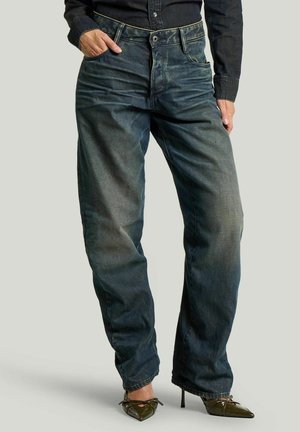 Person trägt lockere dunkelblaue Jeans mit ausgewaschenen Stellen, dunkles Hemd mit Knöpfen und spitze High-Heels, steht mit einer Hand in der Hosentasche.