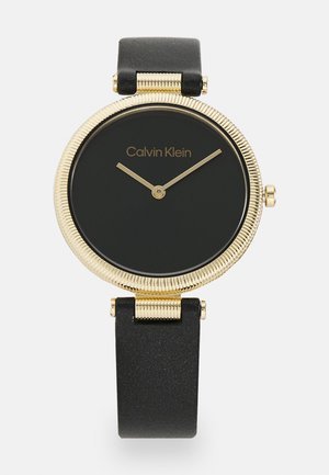 Relógio com bracelete de pele pretas e um bisel texturizado dourado, mostrador preto minimalista e ponteiros dourados finos. Nome da marca visível no mostrador.