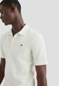 Polo de manga corta en tejido blanco roto, con un cuello abotonado y un pequeño logo marrón en el pecho. Textura suave y diseño ajustado.