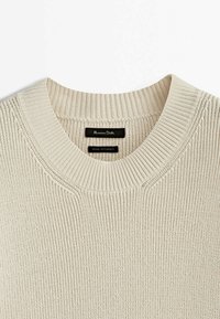 Beżowy sweter dziany z prążkowanym okrągłym dekoltem. Materiał ma fakturowany wzór. Etykiety zawierają informacje o marce i szczegóły dotyczące produkcji.