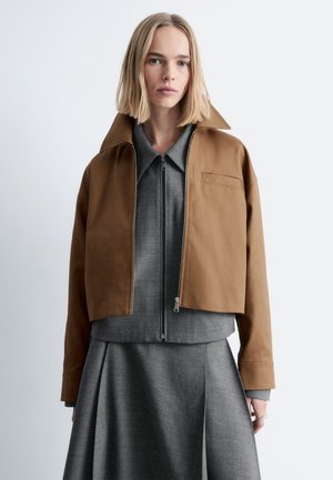 Blonde Frau trägt eine kurze braune Jacke über einem grauen Reißverschluss-Top und einem passenden grauen Rock, steht vor einem schlichten hellen Hintergrund.