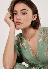 Tenue en tissu vert avec un motif de feuilles dorées, à manches courtes et ceinture à nouer. Collier en chaîne dorée avec un pendentif rectangulaire. Texture lisse.