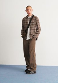 Cămașă cu carouri în nuanțe de maro și crem, tricou alb, pantaloni maro și teniși negri cu dungi albe. Geantă simplă de messenger purtată pe diagonală.