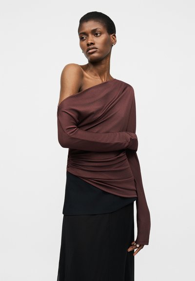Kobieta ubrana w asymetryczną, bordową bluzkę z długim rękawem, z jednym odsłoniętym ramieniem, oraz czarną warstwową spódnicę, stojąca na jednolitym tle.