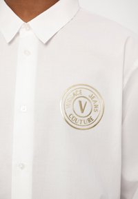 Valge nööpiga särk sileda tekstuuriga, millel on kuldne tikandiga ringikujuline logo, mis ütleb "Versace Jeans Couture."