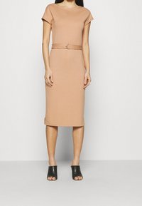 Robe ajustée couleur nude avec manches courtes et taille ceinturée. Tissu doux, longueur au genou, fentes latérales, associée à des mules noires à bouts ouverts.