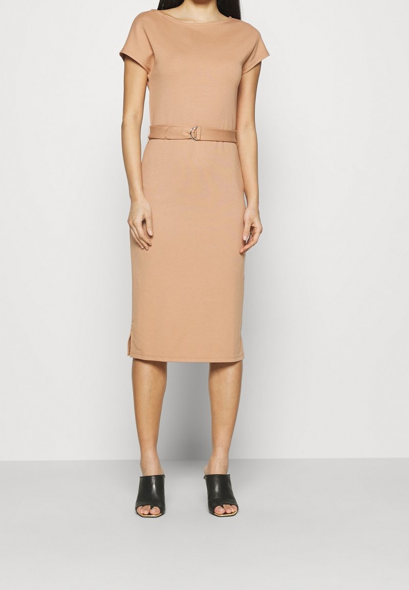 Robe ajustée couleur nude avec manches courtes et taille ceinturée. Tissu doux, longueur au genou, fentes latérales, associée à des mules noires à bouts ouverts.