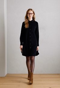 Donna con lunghi capelli che indossa occhiali, un vestito nero con bottoni, calze nere trasparenti e stivali alla caviglia in suede marrone mentre cammina all'interno.