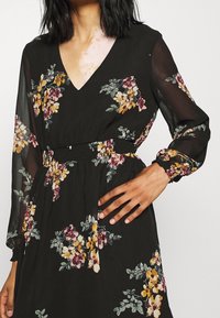Černé květinové šaty s výstřihem do V, elastickým pasem a dlouhými průhlednými rukávy. Vyznačují se květinovými vzory v žluté, růžové a zelené barvě.