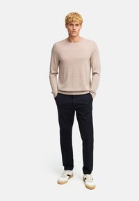 Maglione beige a maniche lunghe con scollo rotondo, abbinato a pantaloni scuri e sneakers bianche con suole beige, in piedi contro uno sfondo neutro.
