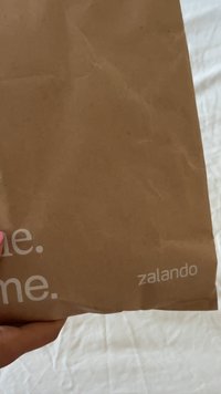 Kraft papirnata shopping torba s lagano nabranom teksturom, s bijelim ispisanim tekstom "me." i "zalando" sprijeda, koju drži ruka.