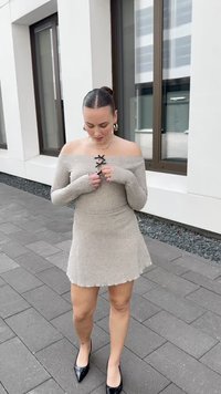 Kvinne justerer sin beige off-shoulder strikkekjole, stående på et brosteinsbelagt fortau ved siden av en moderne bygning med store vinduer.