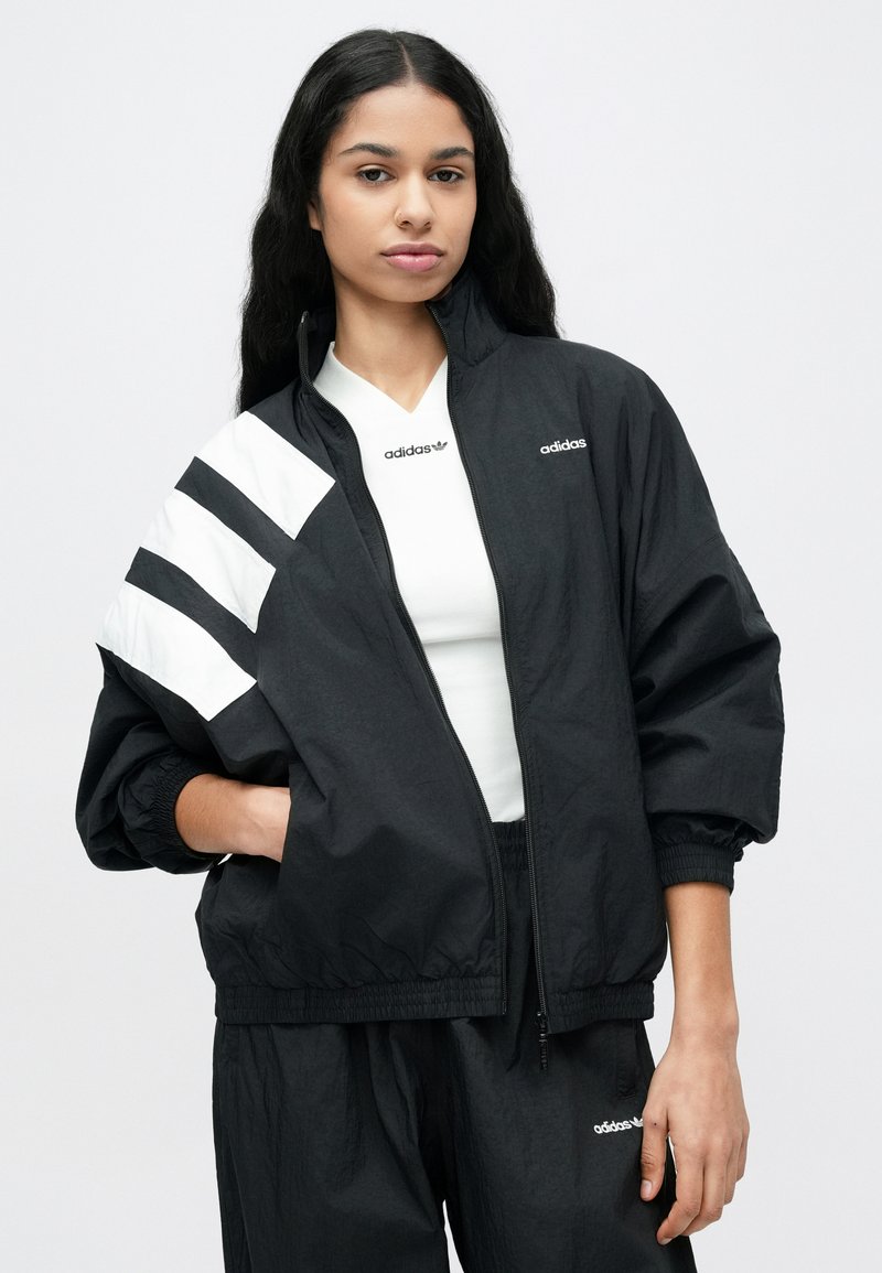 Sort vindjakke med hvide striber på skuldrene, lynlås foran og elastiske manchetter, stylet med en hvid T-shirt med logo under.