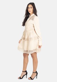 Beiges Kleid mit langen Ärmeln, ausgestattet mit Spitzenbesatz und gestaffeltem Rock. Kombiniert mit schwarzen High-Heel-Sandalen. Glatte Textur, runder Kragen.