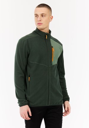 Young man wearing a dark green zip-up fleece jacket with orange zipper details, standing against a white background, looking to the side.