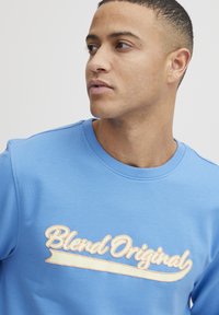 Sudadera azul claro con cuello redondo, que presenta un logotipo "Blend Original" en crema y melocotón. Tejido suave y texturizado. Costuras detalladas.