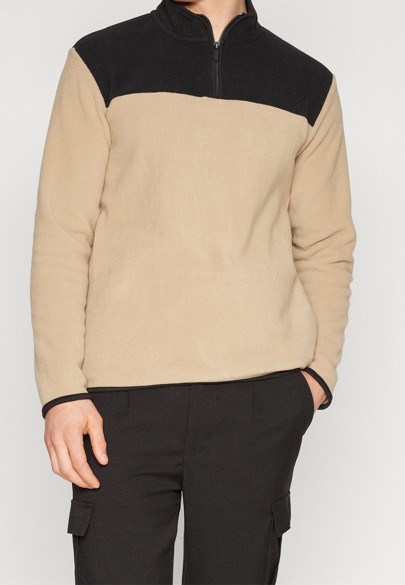 Sudadera de forro polar con cuerpo color beige y cuello negro, que presenta un cierre de media cremallera y tejido texturizado. Con detalles en negro en los puños.