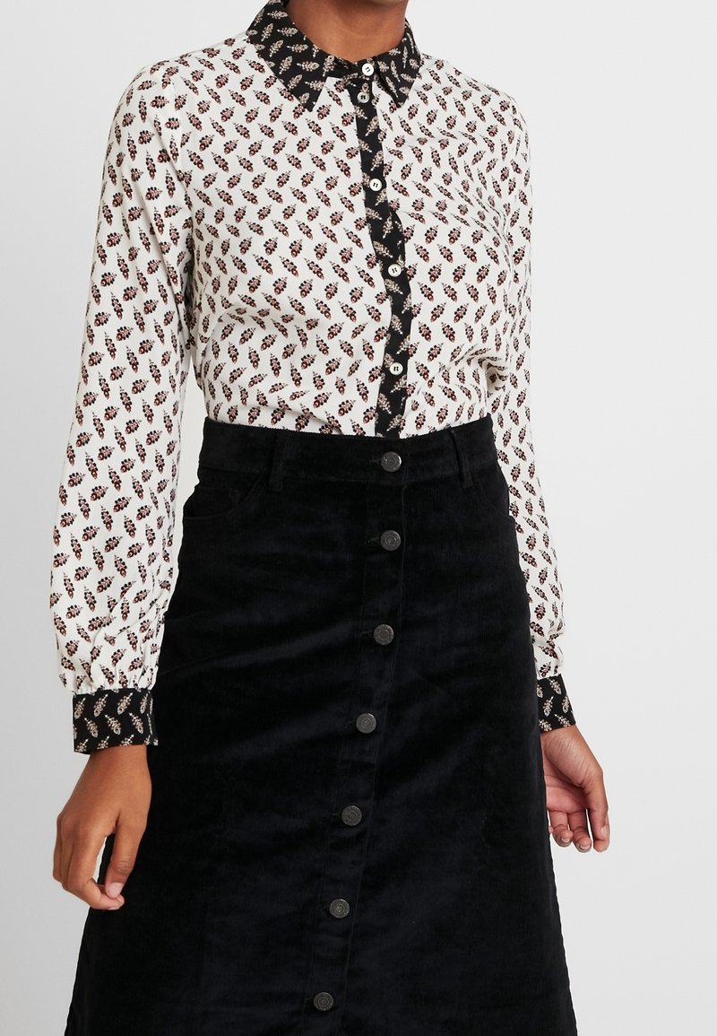 Femme portant un chemisier à manches longues à motifs avec bordure noire et une jupe noire boutonnée.