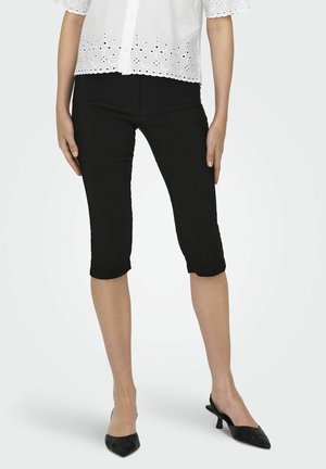 Femme portant un pantalon noir court, un chemisier blanc à petits trous et des talons noirs pointus à bride arrière, debout les bras le long du corps.