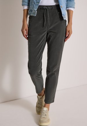 Personne portant un pantalon gris foncé à cordon, des baskets beiges, un haut blanc et une veste en jean bleu clair avec les manches retroussées.