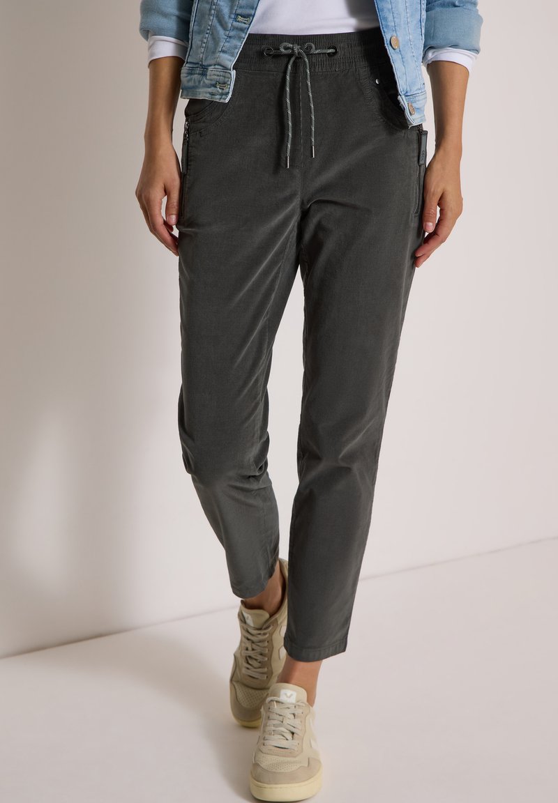 Personne portant un pantalon gris foncé à cordon, des baskets beiges, un haut blanc et une veste en jean bleu clair avec les manches retroussées.