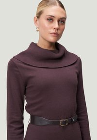 Brauner Pullover mit hohem Rippkragen, langen Ärmeln und taillierter Silhouette, die in der Taille mit einem schwarzen Ledergürtel gerafft ist.