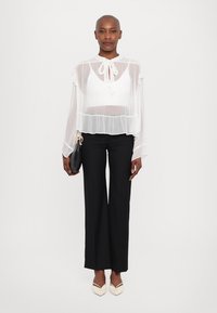Witte, doorzichtige blouse met een striknek, gelaagde ruches, gecombineerd met zwarte, wijde broekspijpen en crèmekleurige puntige hakken. Houdt een zwarte tas vast.