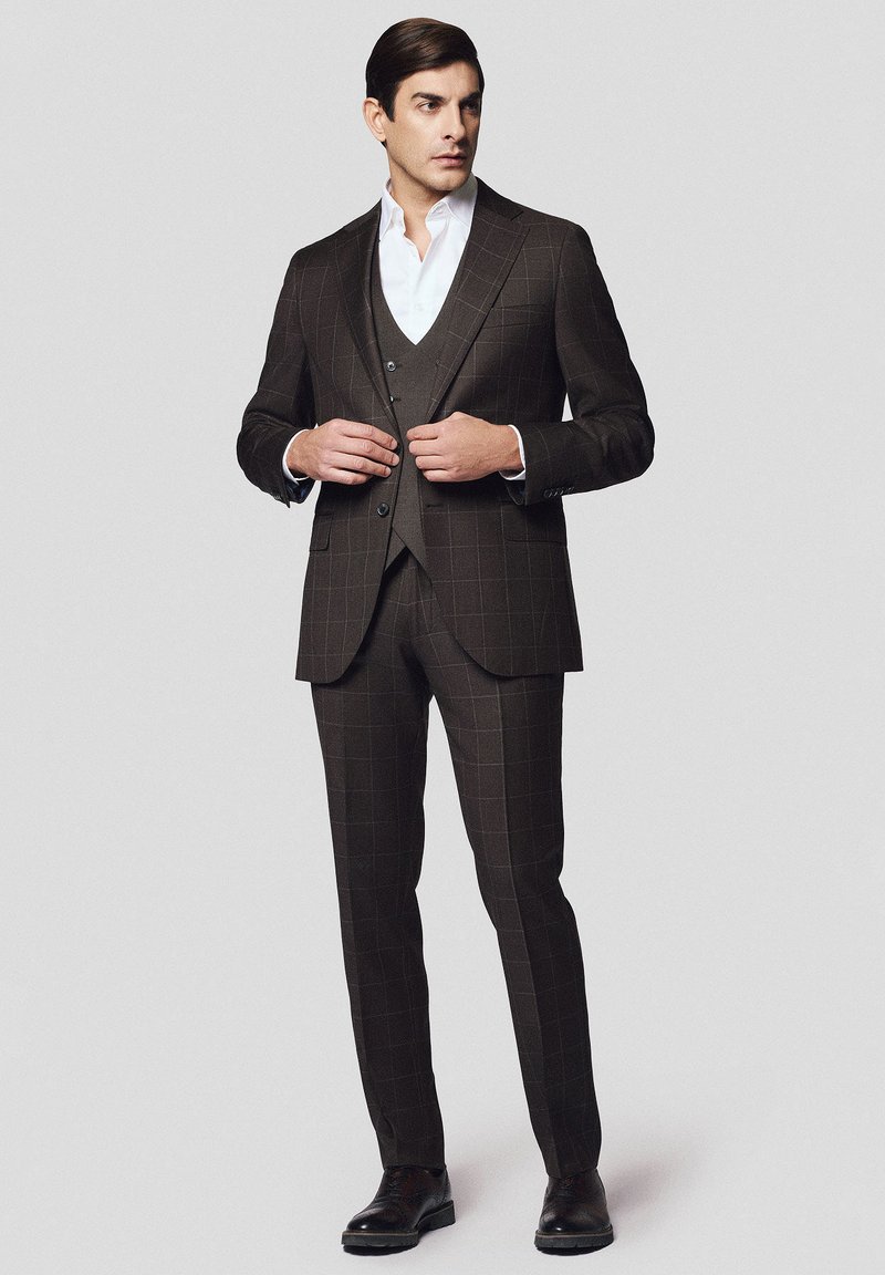 Ciemnobrązowy garnitur trzyczęściowy w kratkę, składający się z dopasowanej marynarki, kamizelki i szytych na miarę spodni. Biała koszula pod spodem, wypolerowane buty.