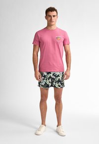 T-shirt di cotone rosa con grafiche "Key West", abbinata a pantaloni da bagno neri con motivi tropicali di foglie e fiori, indossata con sneakers bianche.