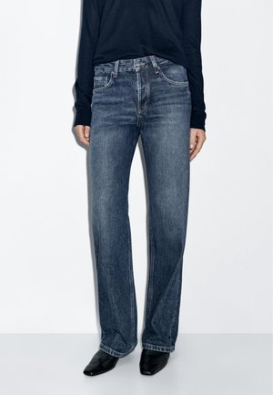 Personne portant un jean droit bleu, des chaussures noires pointues et un haut noir à manches longues, debout devant un fond blanc.