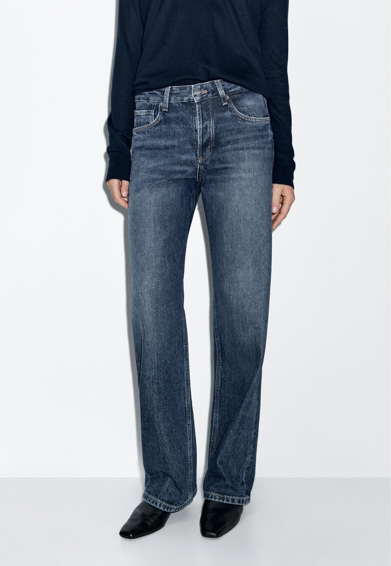 Personne portant un jean droit bleu, des chaussures noires pointues et un haut noir à manches longues, debout devant un fond blanc.