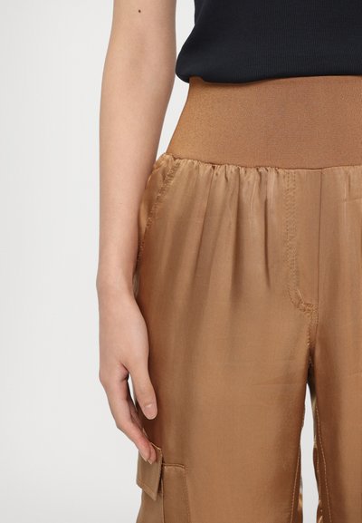 Bruine satijnen broek met een brede elastische tailleband, voorzien van een zijzak en een soepele textuur. Hand rustend op de tailleband.