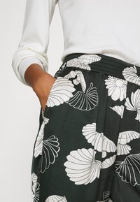 Czarne spodnie z białym wzorem muszli, elastycznym pasem, bocznymi kieszeniami i miękkiego materiału. Modelka ma na sobie białą bluzkę z długim rękawem.