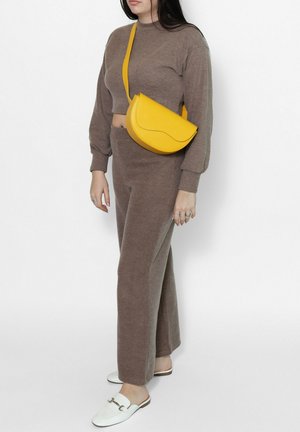 Vrouw met een bruin gebreid kort topje en broek, gele schoudertas en witte instapper loafers, staand tegen een witte achtergrond.