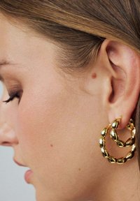 Guldörhängen har en dubbel-lager kedjedesign med släta, ovala länkar. Örhängena är blanka och reflexiva, vilket förstärker deras metalliska finish.