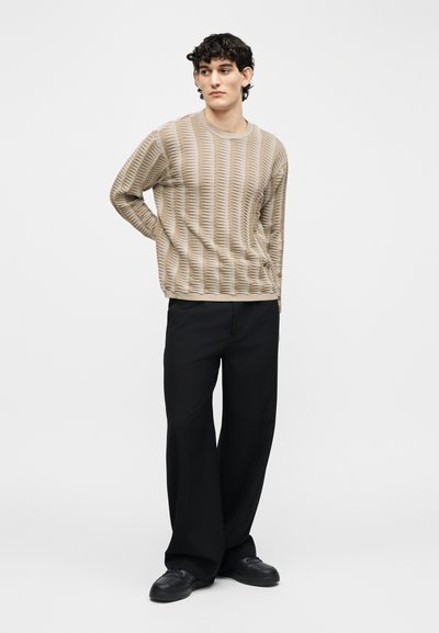 Homme debout, les mains derrière le dos, portant un pull beige texturé, un pantalon large noir et des chaussures noires, sur fond uni.