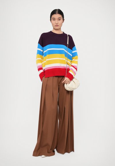 Maglione lavorato a maglia multicolore con righe orizzontali in viola, blu, giallo, rosa e rosso; abbinato a pantaloni a gamba larga marroni a vita alta.