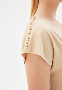 Beige top vyrobený z měkké látky, s v-výstřihem a krajkovými detaily podél ramenního švu, s krátkými rukávy.